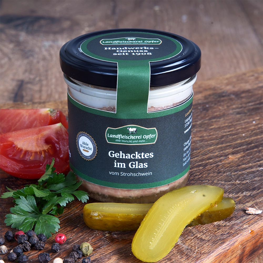 Ein Glas Gehacktes mit schwarzem Schraubverschluss und schwarzem Etikett steht auf einem Holzbrett. Daneben liegen Gewürzgurken und Tomaten.