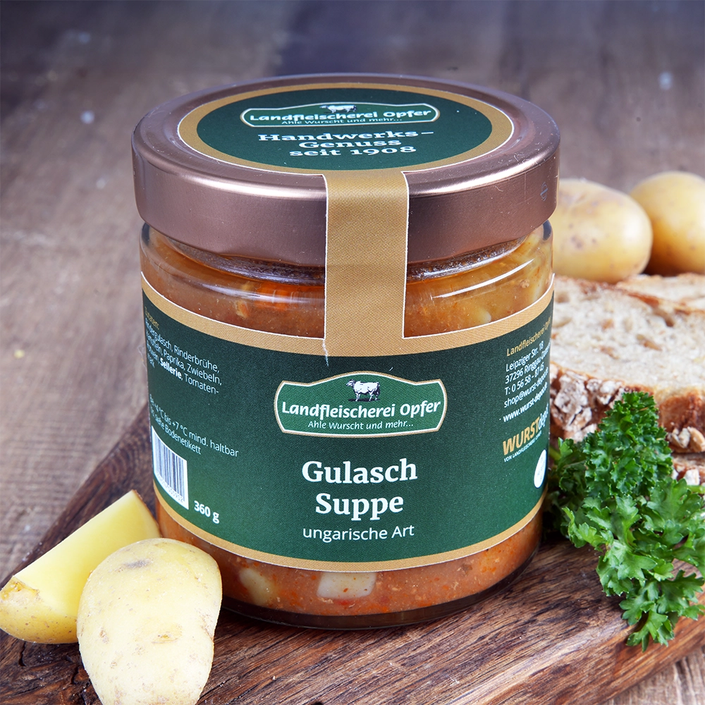 Ein Glas Gulaschsuppe mit Schraubverschluss und grünem Etikett steht auf einem Holzbrett. Daneben liegen Kartoffeln, Petersilie und Brotscheiben.