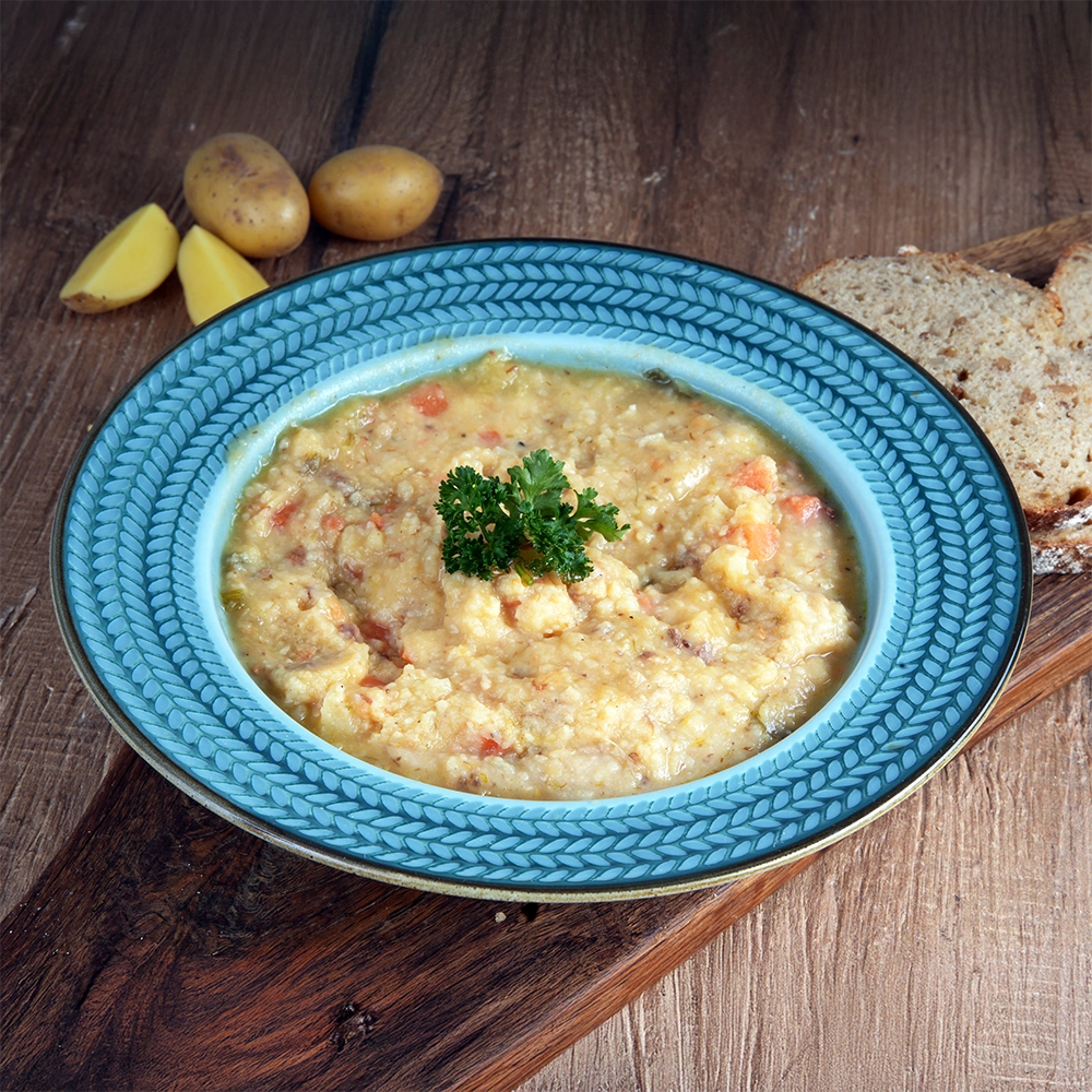 Ein blauer Teller mit Ornamenten ist gefüllt mit Kartoffelsuppe und steht auf einem Holzbrett. Daneben liegen Petersilie und Kartoffeln.