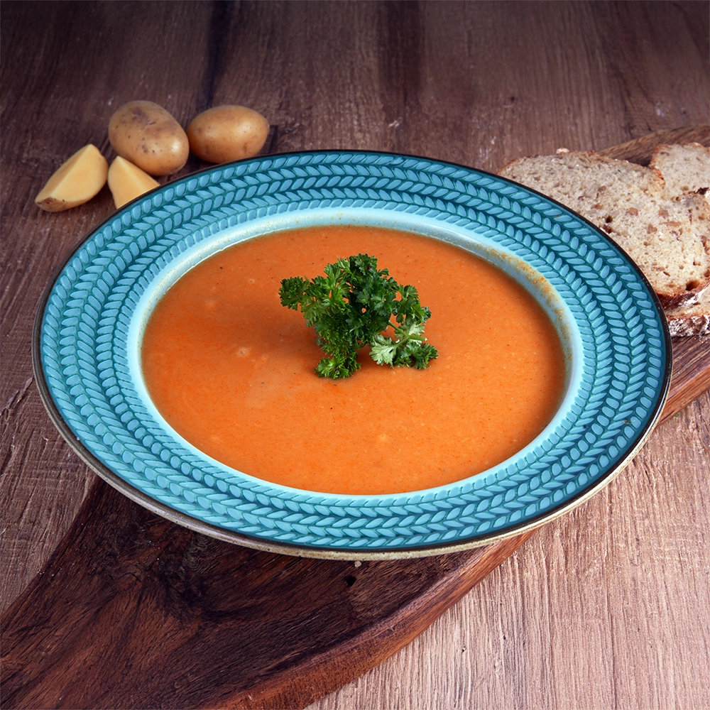 Ein blauer Teller mit Ornamenten ist gefüllt mit Kürbissuppe und steht auf einem Holzbrett. Daneben liegen Kartoffeln und Brotscheiben.