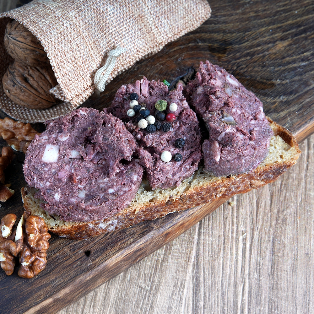 Eine Brotscheibe mit Rotwurst liegt auf einem Holzbrett. Daneben liegen Walnüsse.