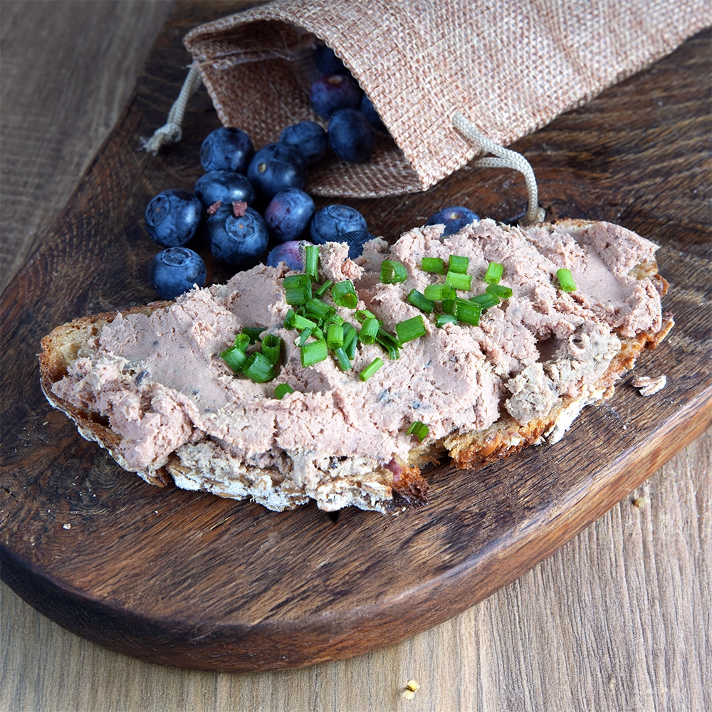 Eine Brotscheibe mit Waldleberwurst und Schnittlauch liegt auf einem Holzbrett. Daneben liegen Heidelbeeren.