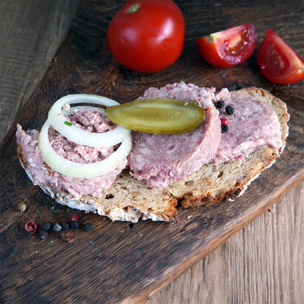 Eine Brotscheibe mit Wildschweinsülze, garniert mit Zwiebelringen und Essiggurke liegt auf einem Holzbrett. Daneben liegen geschnittene Tomaten.